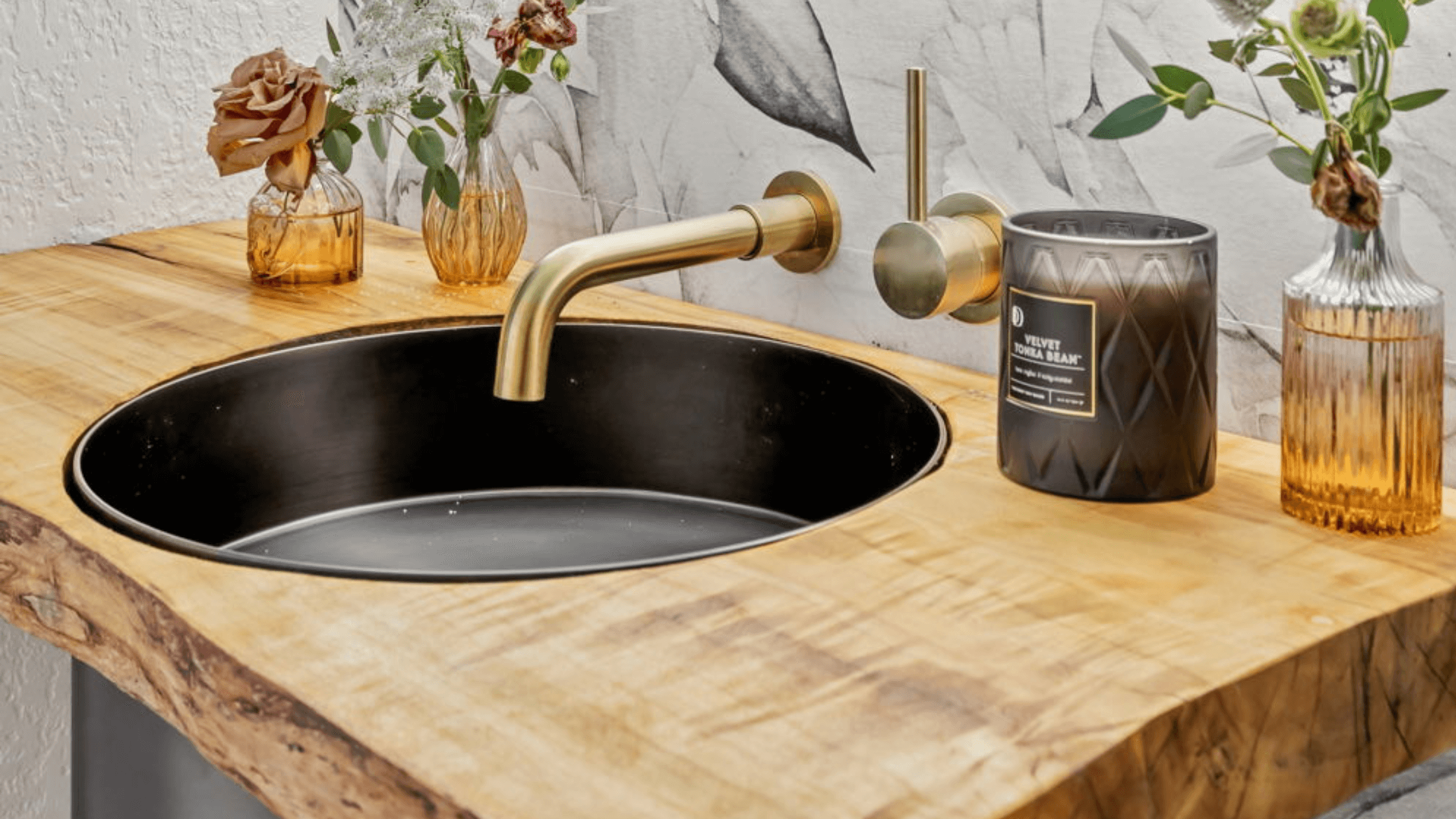 Wall-mounted gold faucet with round black sink set in a live-edge wood vanity, styled with amber glass vases and a luxury candle — modern rustic bathroom design by Wrecked and Refined.